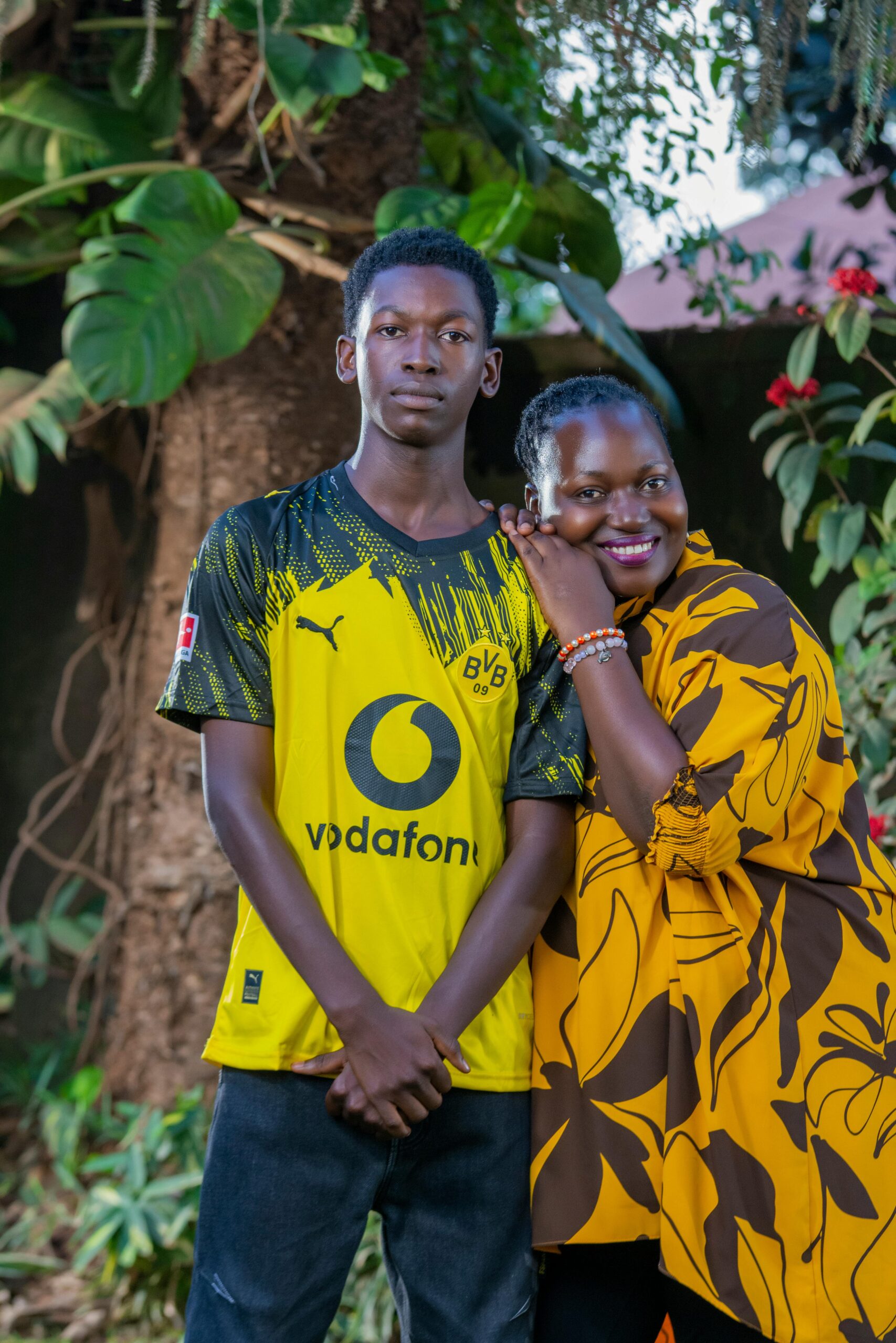 Home A joyful mother and son pose together outdoors in colorful outfits, surrounded by lush greenery.